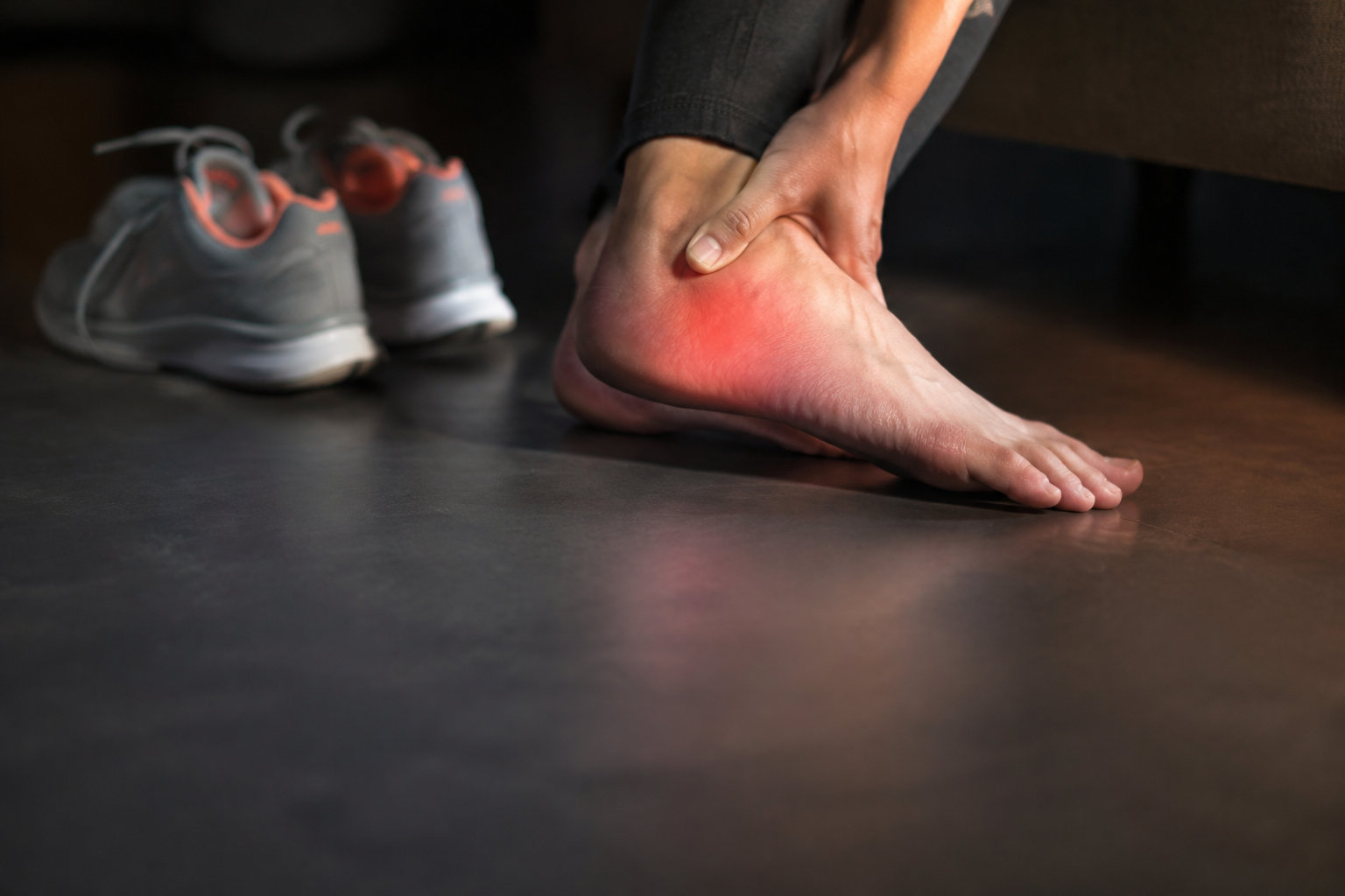 Personne se tenant le talon douloureux avec une zone rouge indiquant une inflammation, chaussures de sport en arrière-plan.